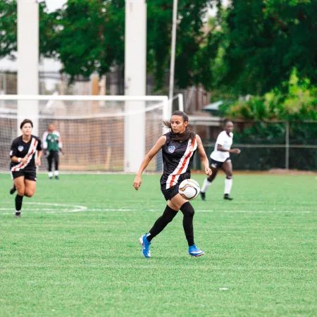 Female soccer player in black uniform controlling the ball on green field with players and goal in background