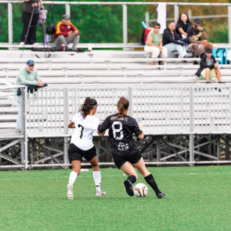Two female soccer players competing for the ball on a green field with spectators in the background.
