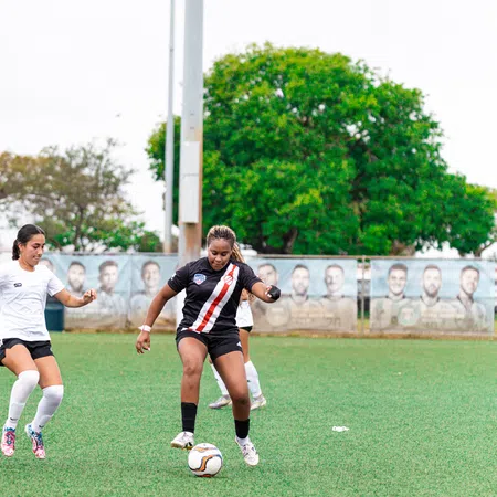 Two female soccer players competing for the ball on a green field with trees and banners in the background
