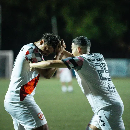 Two soccer players in white uniforms celebrate by bumping heads on a grassy field during a night game.
