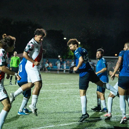Night soccer match showing a player heading the ball as defenders and teammates watch on grassy field