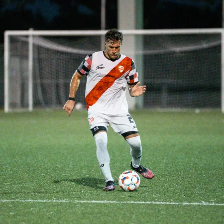 Soccer player in white and red uniform controlling the ball on a green field with goalpost in the background at night