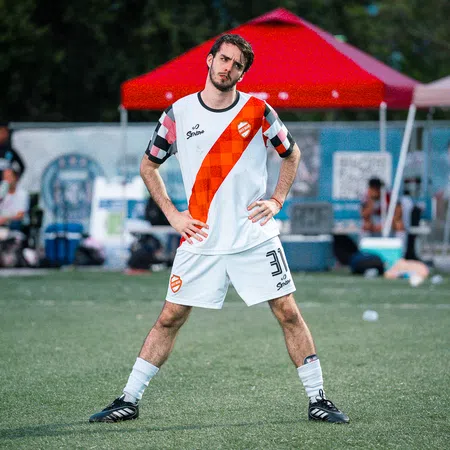 Male soccer player in white and orange uniform standing on green field with hands on hips under red canopy tent.