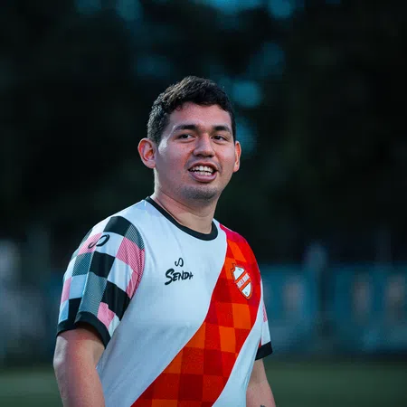 Young male soccer player in a checkered and red jersey smiling on a field during evening practice.