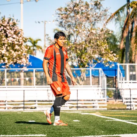 Soccer player in red and black uniform walking on field with palm trees and benches in background under clear sky