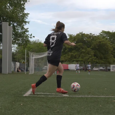 Female soccer player in black jersey preparing to kick ball near goal on grassy field during game.