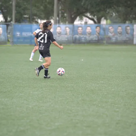 Female soccer player in black jersey controlling the ball on a green field against opponents in white jerseys.