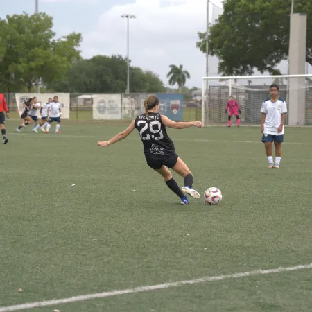 Female soccer player in black jersey kicking ball on outdoor field during game with opponents and referee.