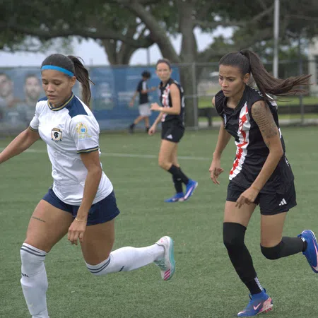 Female soccer players in action on a green field during a daytime match with trees and a fence in the background