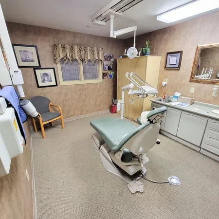 Dental clinic room with a green dental chair, overhead light, wooden cabinets, and framed pictures on beige walls.