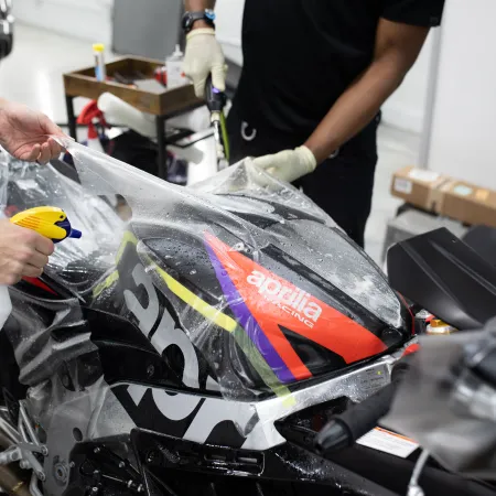 Two people applying protective clear film on Aprilia racing motorcycle in a workshop setting
