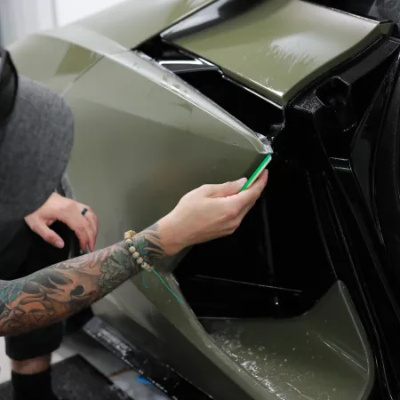 Tattooed person applies protective film to sleek green car hood using a green tool in a workshop.