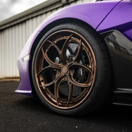Close-up of a purple sports car's bronze alloy wheel and tire on asphalt near a corrugated metal wall.