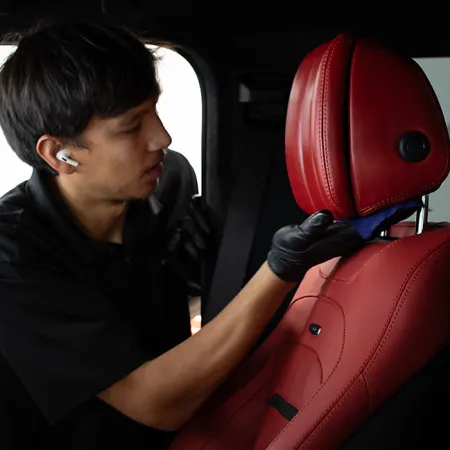 Technician cleaning and detailing red leather car seat inside vehicle with a microfiber cloth and gloves