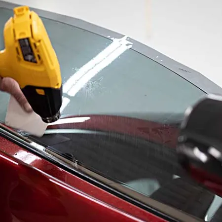 Person using a yellow heat gun to remove window tint film from a red car window indoors.