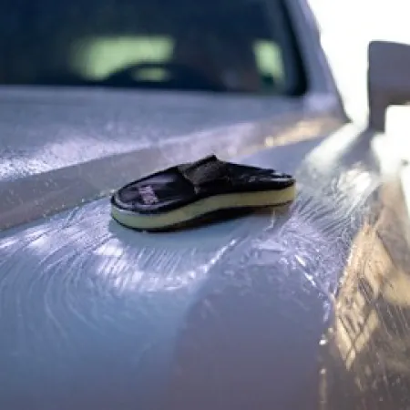 Foam applicator pad on a wet car hood during washing, showing soap and water streaks under sunlight.