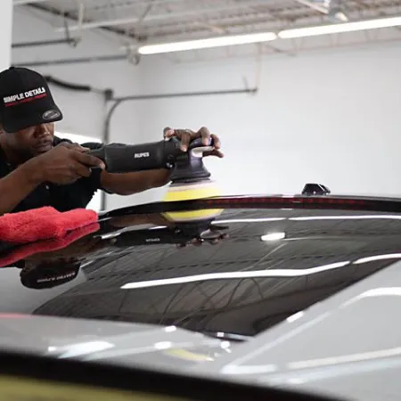 Man using a polishing machine to detail a glossy black car roof in a bright garage.