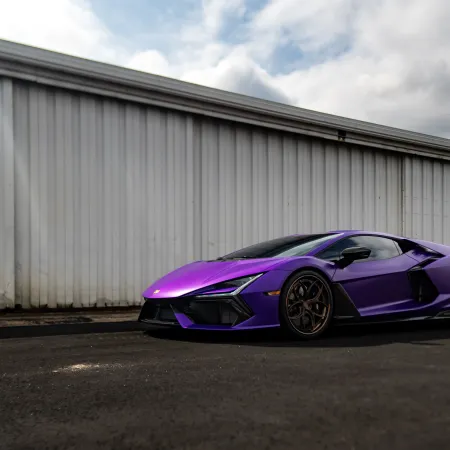 Purple sports car parked on asphalt in front of a large white industrial building with a windy orange windsock.