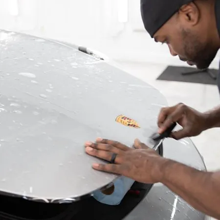 Man applying protective film to the hood of a silver Porsche car in a well-lit garage.