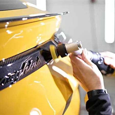Close-up of hands polishing the rear carbon fiber detail on a yellow sports car with an electric buffer.
