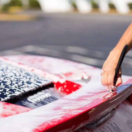 Person washing a red car with soap suds in an outdoor parking lot on a sunny day.