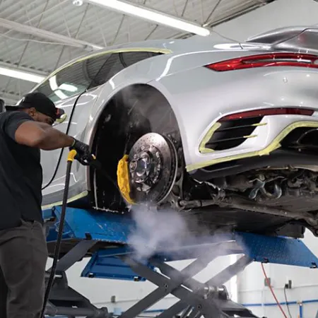 Technician steam cleans brake disc of silver sports car elevated in a bright automotive workshop.