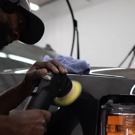 Man using a polishing machine to buff a black car's front panel near the headlight in a workshop.