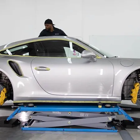 Silver sports car on a hydraulic lift without wheels in a garage with a mechanic in the background