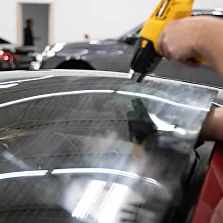 Person installing window tint film on a sports car windshield using a heat gun in a garage.