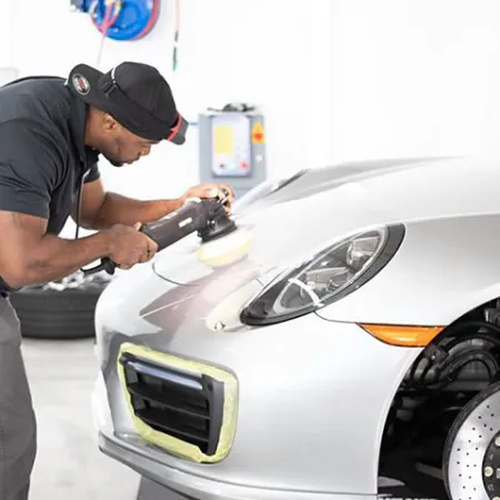 Man polishing the front of a silver sports car with a buffer machine in a bright garage.
