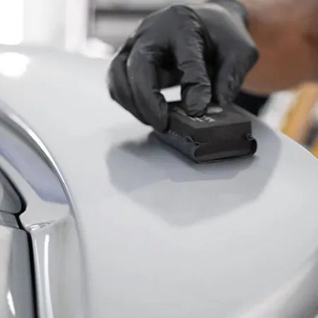 Person wearing black gloves polishing a white car surface using a black polishing tool indoors.
