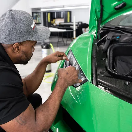 Man wearing gray cap detailing the headlight of a bright green sports car in a garage.