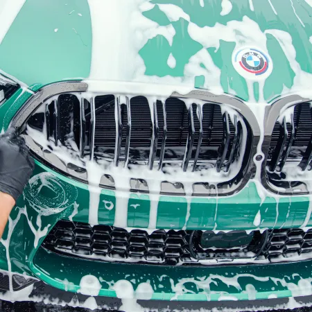 Close-up of a green BMW car front grille being cleaned with soap foam by a worker wearing black gloves.