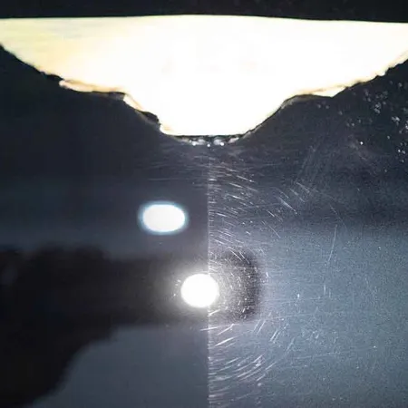 Reflection of hand holding phone capturing light spots on a dark shiny surface with scratches and smudges.