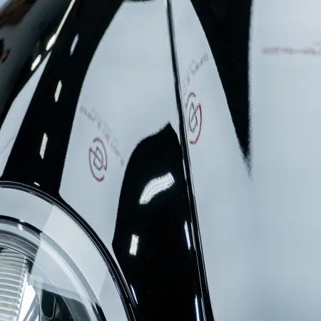 Close-up of a shiny black car headlight and hood reflecting white ceiling lights and logos on a showroom floor.