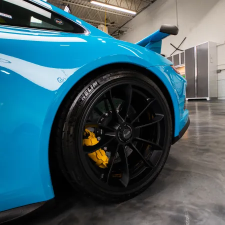 Close-up of a blue sports car’s rear wheel with black rims and yellow brake calipers in a garage.