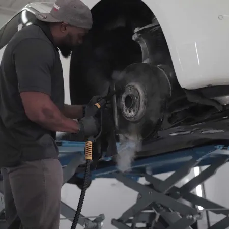 Mechanic using steam cleaner to clean car brake assembly on a lifted white vehicle in a garage.