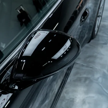 Close-up of sleek black car side mirror reflecting overhead lights on shiny surface.