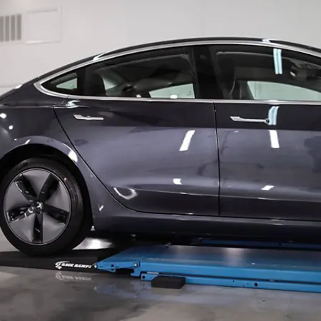Dark gray electric sedan on blue inspection platform in a clean automotive workshop with reflective floor.