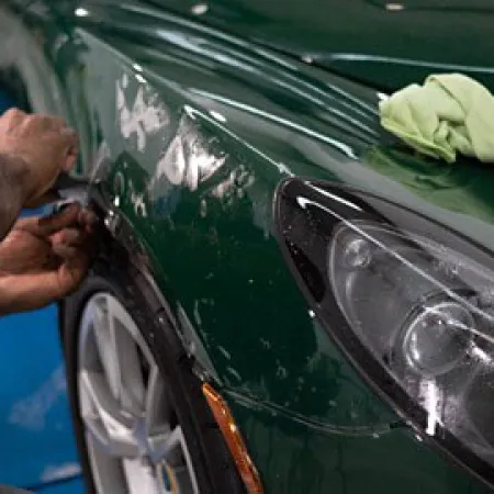 Person repairing damage on a dark green car near the front headlight and wheel in a workshop.