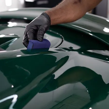 Person polishing a glossy dark green car surface with a blue sponge wearing black gloves indoors.
