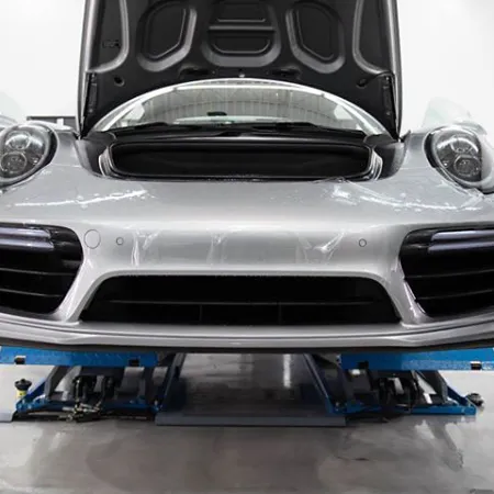 Silver sports car with hood open on lift in garage, mechanic inspecting the front bumper closely.