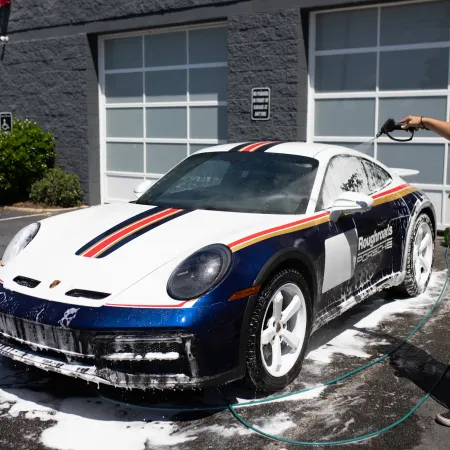 Person washing a white and blue sports car with foam and water outside a garage on a sunny day