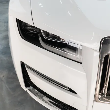 Close-up of a white luxury car's front, featuring a sleek headlight and chrome grille on a polished concrete floor.