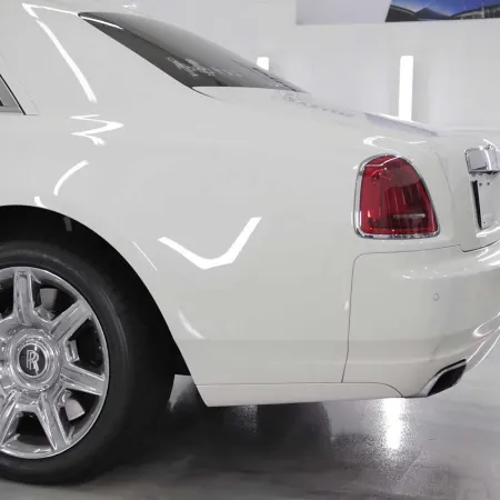 Rear side of a white luxury Rolls-Royce car showcasing chrome wheel and tail light in a bright showroom.