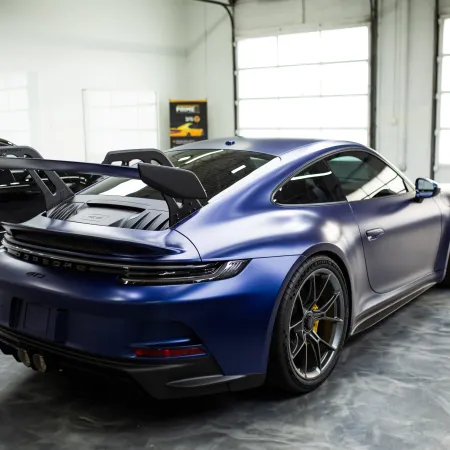 Matte blue Porsche 911 GT3 with large rear wing parked inside a bright garage with concrete floor.