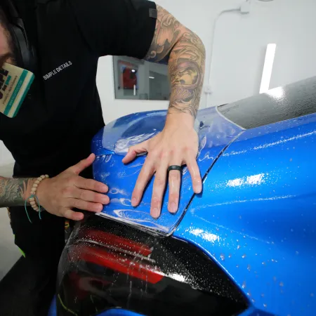 Tattooed man applies protective film to blue car's rear panel in a detailing workshop.