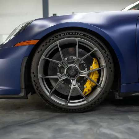 Close-up of a matte blue sports car front wheel with yellow brake caliper and Michelin tire in a garage.