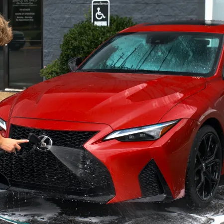 Young man washing a red sports sedan outside on a sunny day with soap and water spray.