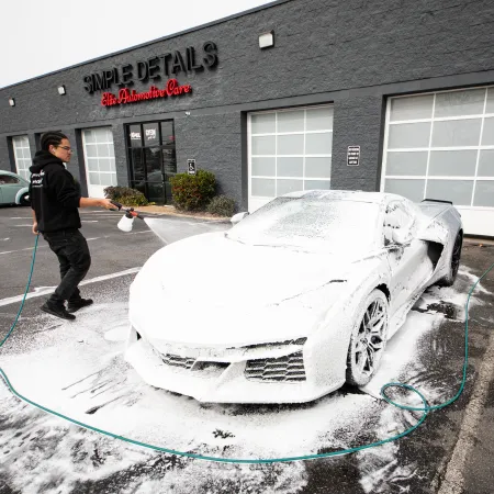 Man washing a luxury sports car covered in foam outside Simple Details Auto Care on a cloudy day.